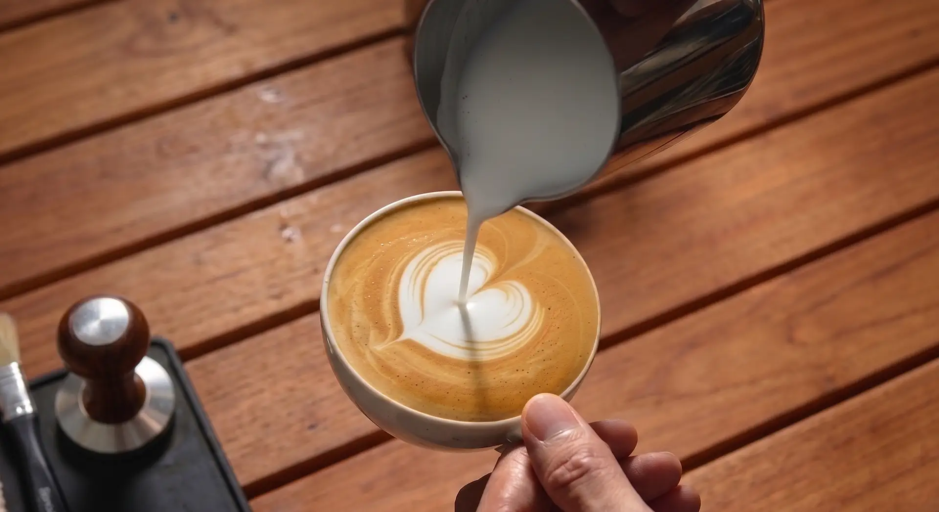 Eine Hand gießt Milch aus einem Kännchen in eine Kaffeetasse und formt ein Herz in der Crema; links daneben liegt Barista-Zubehör auf einem Holztisch.