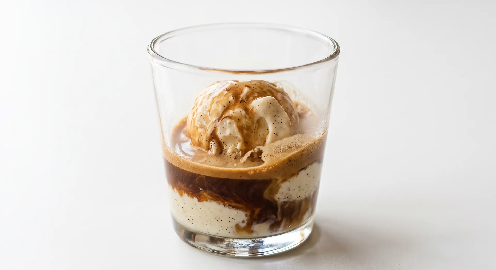 Ein transparentes Glas, das mit Schichten von dunkelbraunem Espresso, cremefarbener, flüssigerer Eiskrem und einer scoopfesten, beigefarbenen Kugel Eis gefüllt ist.