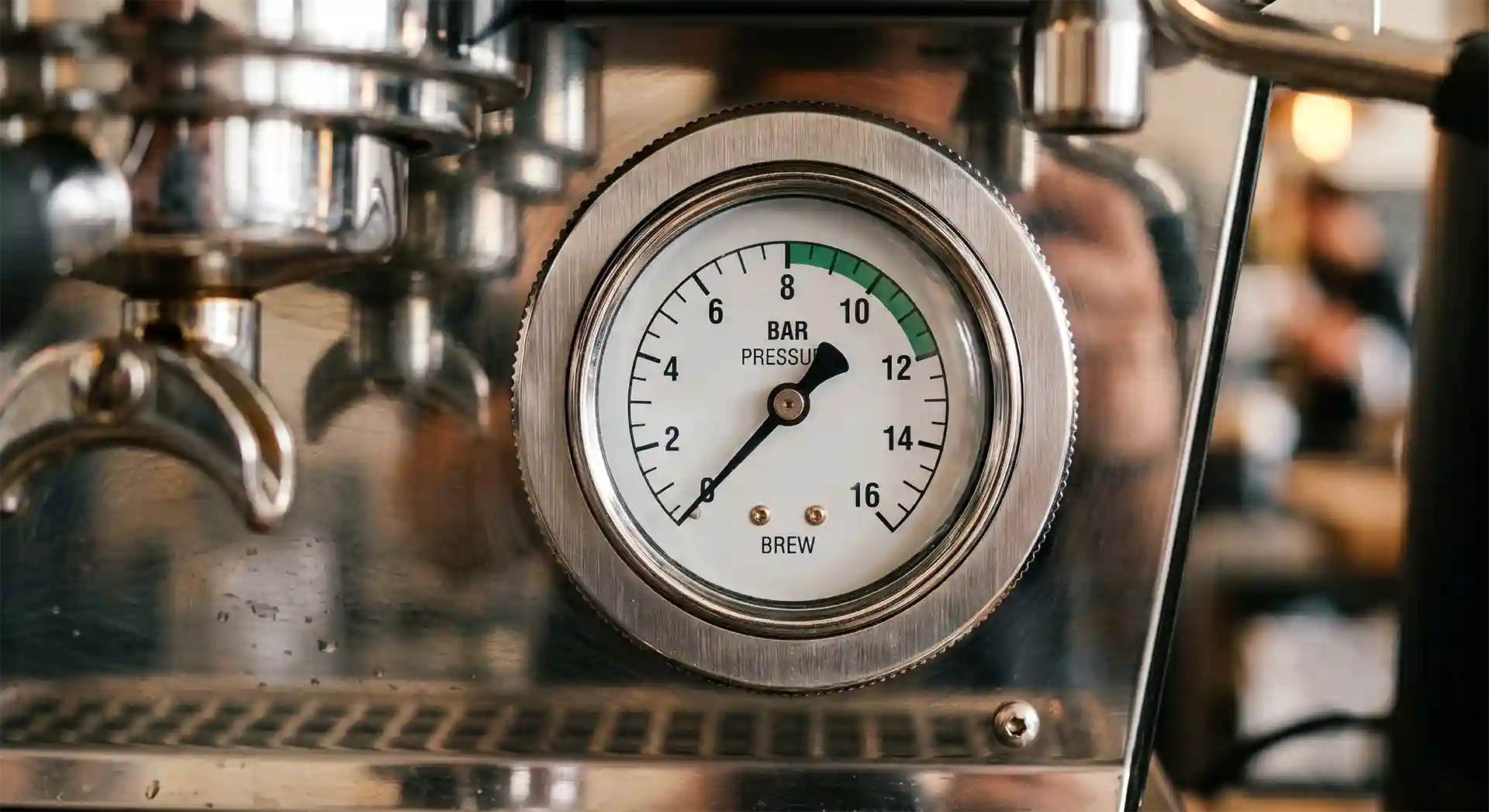 Eine Nahaufnahme eines runden Druckmanometers an einer glänzenden, sauberen Espressomaschine aus Edelstahl. Der Zeiger der Anzeige steht auf Null Bar. Im Hintergrund ist der Siebträger der Maschine leicht unscharf zu sehen.